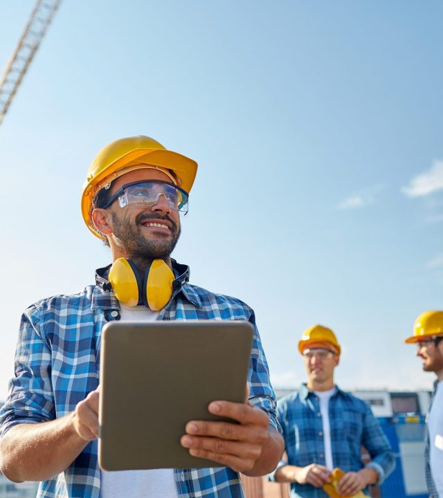 business, building, industry, technology and people concept - smiling builder in hardhat with tablet pc computer over group of builders at construction site